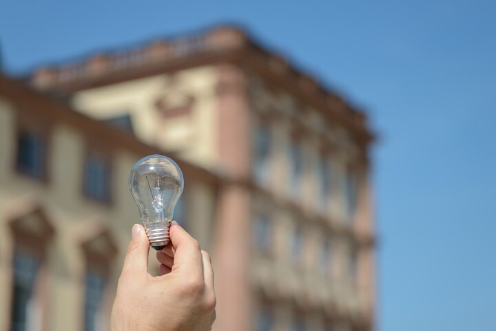 Eine Hand hält eine Glühlampe. Im Hintergrund befinden sich Teile des Barockschlosses Mannheim.