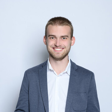 Portrait von Herrn Spils. Er hat kurze blonde Haare, einen Bart und trägt über einem weißen Hemd ein grau-blaues Jackett.