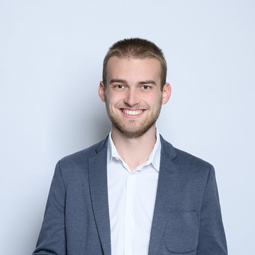 Portrait von Herrn Spils. Er hat kurze blonde Haare, einen Bart und trägt über einem weißen Hemd ein grau-blaues Jackett.