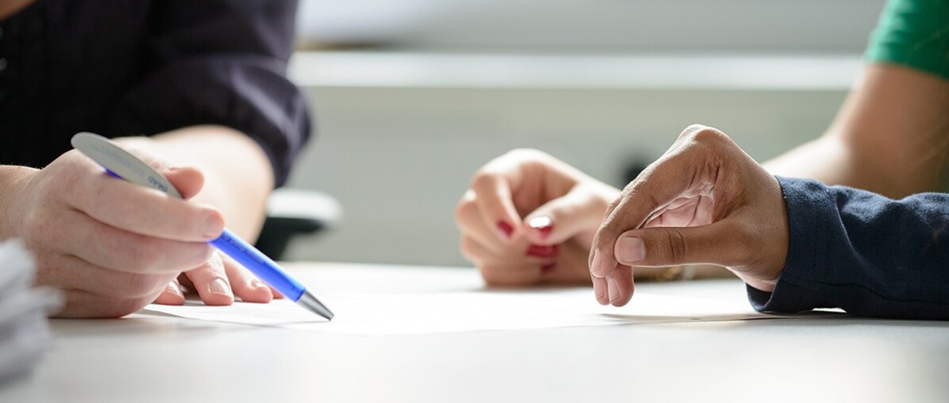 Drei Personen legen einen Arm auf einen Tisch. Eine Person die ein dunkles Oberteil trägt zeigt mit der anderen Hand mit einem dunkelblauen Kugelschreiber etwas auf einem Blatt. Eine der anderen Personen hat rot lackierte Fingernägel und trägt ein dunkelgrünes T-Shirt. Die andere Person trägt ein langes dunkles Oberteil.