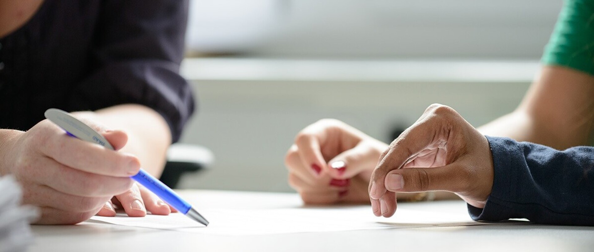 Drei Personen legen einen Arm auf einen Tisch. Eine Person, die ein dunkles Oberteil trägt, zeigt mit der anderen Hand mit einem dunkelblauen Kugelschreiber etwas auf einem Blatt. Eine der anderen Personen hat rot lackierte Fingernägel und trägt ein dunkelgrünes T-Shirt. Die andere Person trägt ein langes dunkles Oberteil.