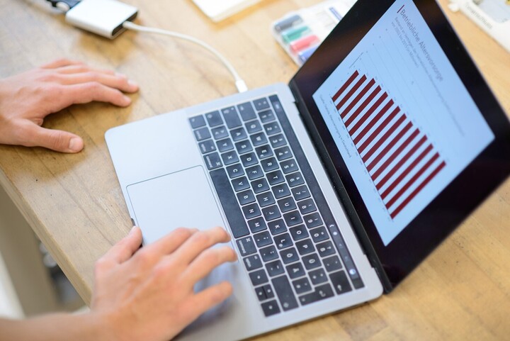 Ein aufgeklappter Laptop auf einem Tisch, links zwei Hände.
