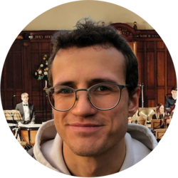 Portrait von Felix, einem Bachelorstudenten der Volkswirtschaftslehre. Er hat kurze braune Haare, trägt eine Brille und einen grauen Hoodie. Er steht vor einem Orchester.