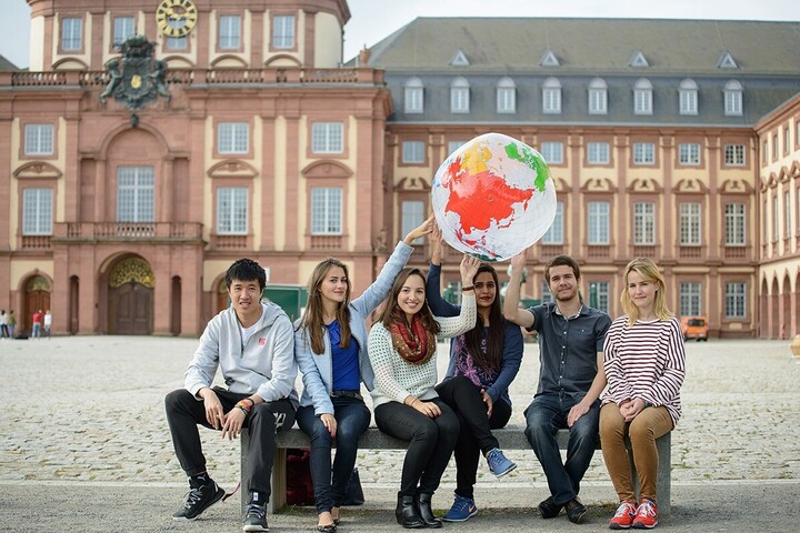 Sechs Studierende sitzen auf einer Bank vor der Universität Mannheim. Einige von ihnen halten gemeinsam eine Weltkugel aus Plastik über ihren Köpfen. 