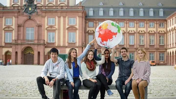 Sechs Studierende sitzen auf einer Bank vor der Universität Mannheim. Einige von ihnen halten gemeinsam eine Weltkugel aus Plastik über ihren Köpfen.