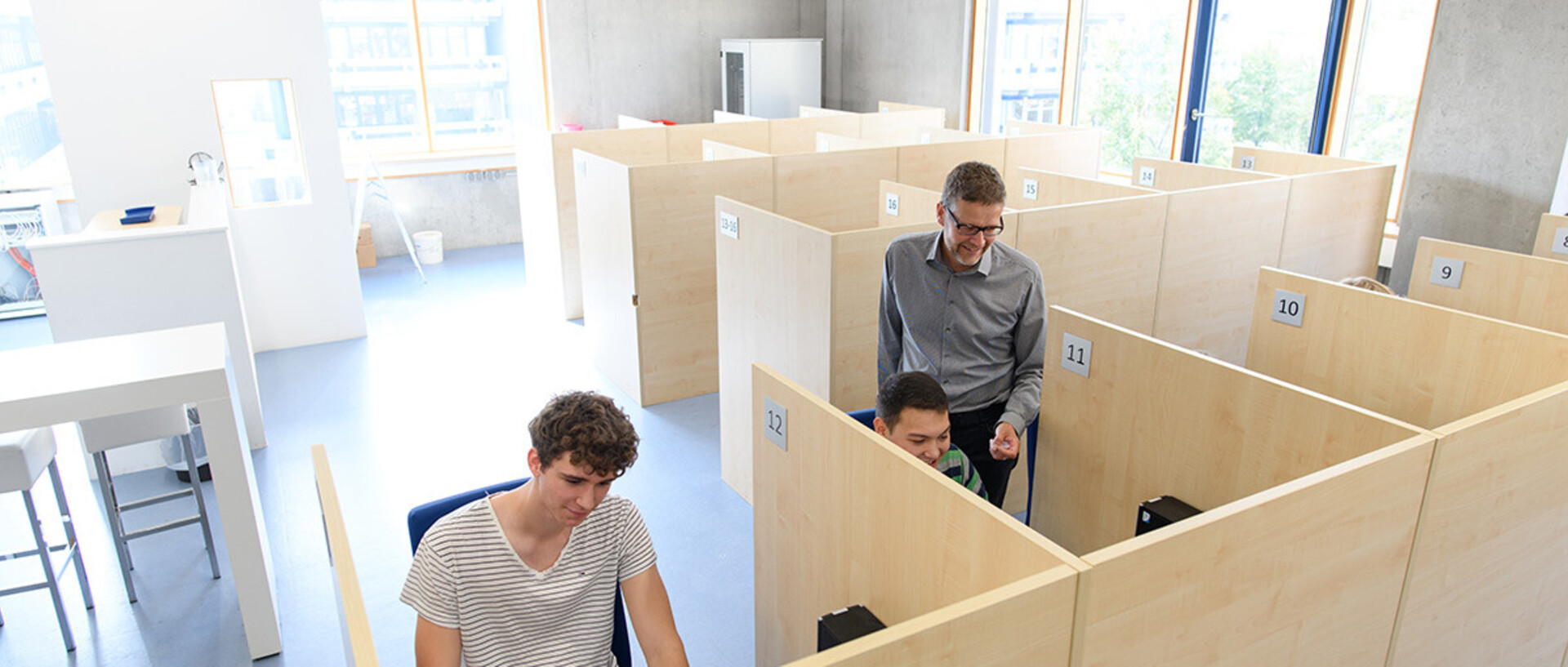 Ein heller Raum mit mehreren Arbeitskabinen aus Holz. Zwei Personen sitzen in separaten Kabinen, Professor Orzen Person steht daneben. Große Fenster im Hintergrund lassen Tageslicht herein.