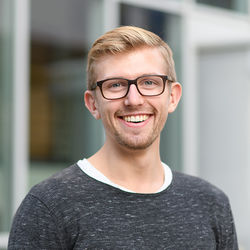 Portrait von Sven, einem Studenten der Volkswirtschaftslehre. Er hat blonde kurze Haare, trägt Vollbart und Brille. Sven trägt ein weißes Oberteil und darüber einen dunkelgrauen Pullover. Sven lächelt in die Kamera.