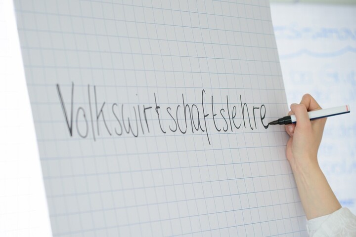 Auf ein Flipchart, welches mit kariertem Papier bespannt ist, wird mit einem schwarzen Filzstift „Volkswirtschaftslehre“ geschrieben.