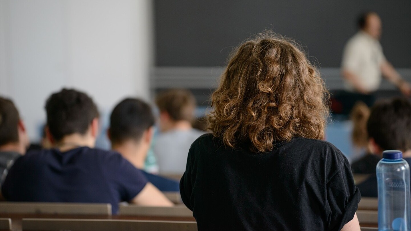 Aufnahme von Hinten in einem Hörsaal. Der Fokus liegt auf einer Studentin mit braunen lockigen Haaren und einem schwarzen Oberteil. Im Hintergrund sitzen weitere Studierende.