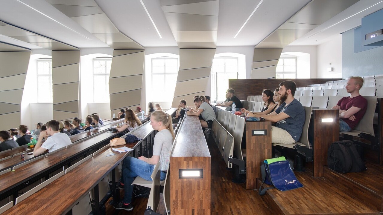 Aufnahme aus dem Hörsaal SO 108 im Ostflügel der Universität Mannheim. In dem Hörsaal sitzen viele Studierende. Die Wände des Hörsaals sind weiß mit grauen Elementen, die Tische sind dunkelbraun.