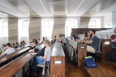 Aufnahme aus dem Hörsaal SO 108 im Ostflügel der Universität Mannheim. In dem Hörsaal sitzen viele Studierende. Die Wände des Hörsaals sind weiß mit grauen Elementen, die Tische sind dunkelbraun.