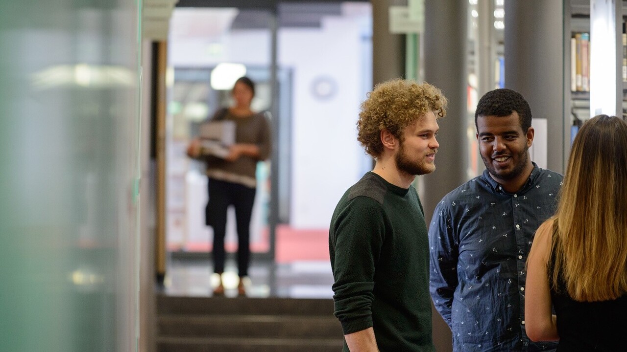 Auf der rechten Bildseite stehen drei Studierende und unterhalten sich. Auf der linken Bildseite steht im Hintergrund eine Person.