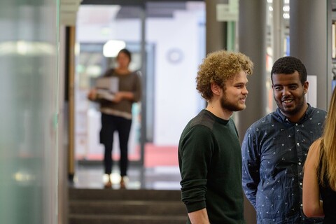 Auf der rechten Bildseite stehen drei Studierende und unterhalten sich. Auf der linken Bildseite steht im Hintergrund eine Person. 