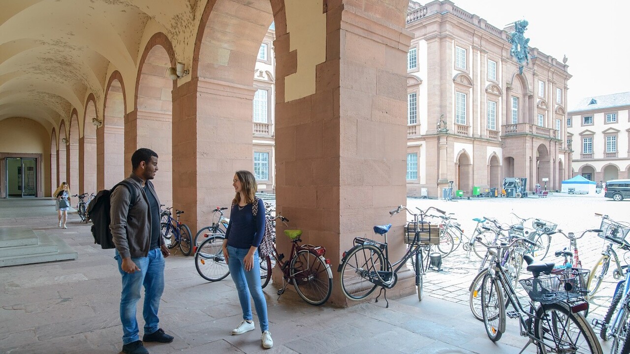 Eine Studentin und ein Student stehen in einem Gang der linksseitig an den Ehrenhof grenzt und unterhalten sich. In den Bögen des Gangs stehen viele Fahrräder.