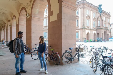 Eine Studentin und ein Student stehen in einem Gang der linksseitig an den Ehrenhof grenzt und unterhalten sich. In den Bögen des Gangs stehen viele Fahrräder.