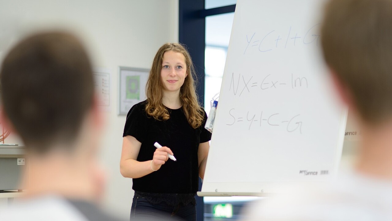 Eine Studentin steht an einem Flipchart. Sie hält einen geöffneten Filzstift in der Hand und blickt in Richtung der Zuhörenden. An den beiden Bildseiten erkennt man zwei Köpfe.