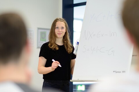 Eine Studentin steht an einem Flipchart. Sie hält einen geöffneten Filzstift in der Hand und blickt in Richtung der Zuhörenden. An den beiden Bildseiten erkennt man zwei Köpfe. 