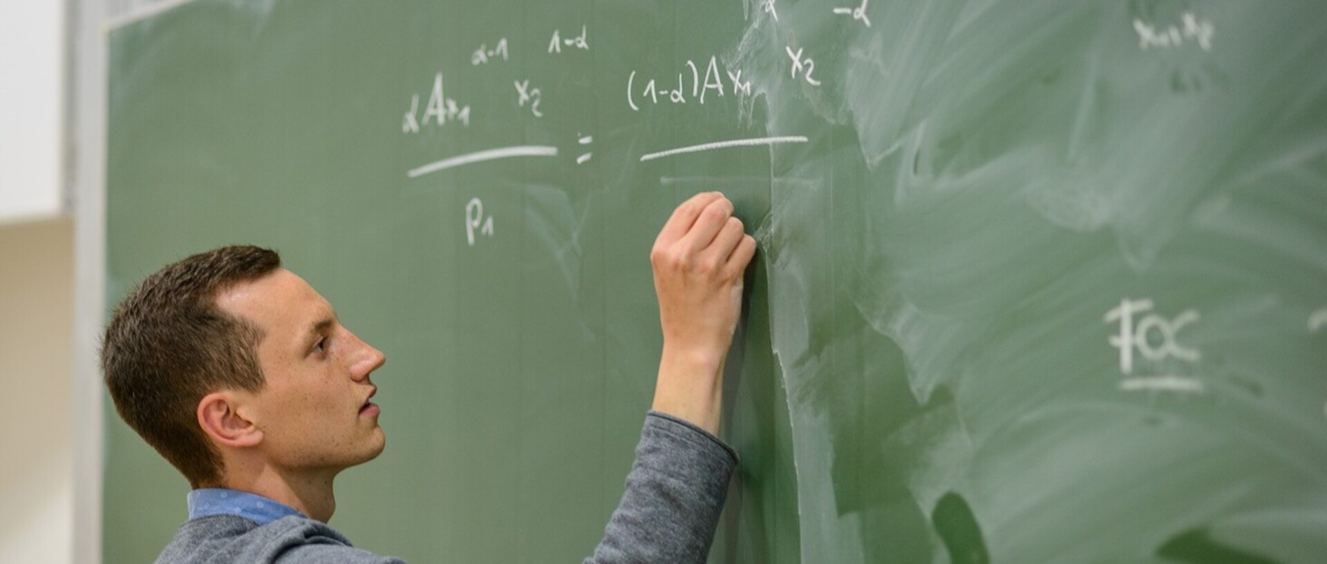 Ein Student beschreibt eine grüne Tafel mit weißer Kreide. Er hat kurze dunkelbraune Haare und trägt ein blaues Hemd mit weißen Punkten und darüber ein graues Oberteil.