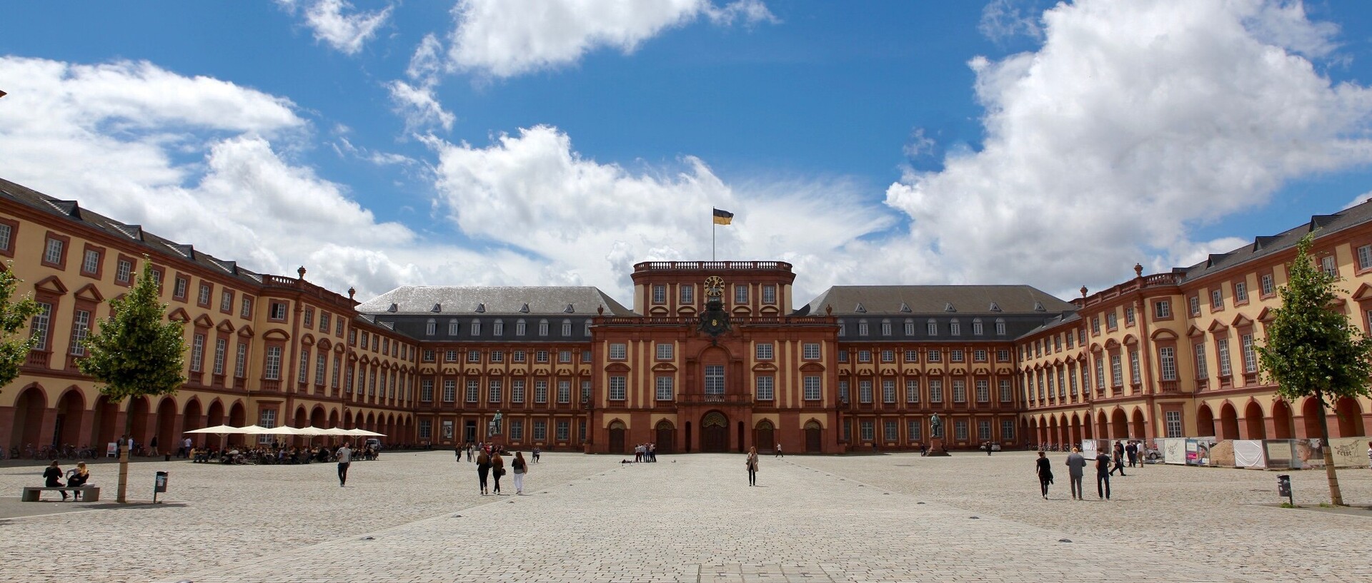 Blick vom Tor des Ehrenhofs über den Ehrenhof auf die Universität Mannheim. Auf dem Ehrenhof stehen am linken und rechten Bildrand Bäume, und ein paar Personen flanieren über den Platz.
