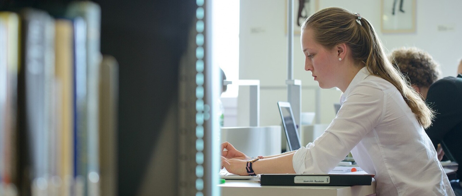 A student with long blonde hair and a white blouse is sitting at a table in the Ehrenhof library.