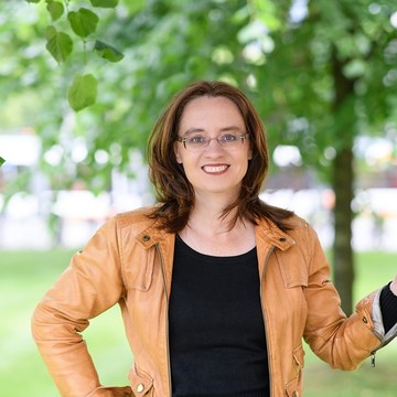 Eine Wissenschaftlerin trägt eine Brille und ihr braunes Haar offen. Über ihrem schwarzen Oberteil trägt sie eine hellbraune Lederjacke und lehnt ihre linke Hand an einen Baum.