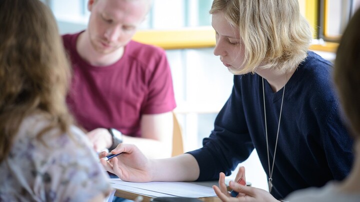 Eine Studentin mit schulterlangen blonden Haaren, welche ein blaues Shirt trägt, sitzt an einem Tisch und füllt ein Blatt aus. Im Hintergrund sitzt ein Student mit blonden kurzen Haaren und einem roten Shirt.
