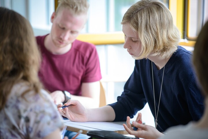 Eine Studentin mit schulterlangen blonden Haaren, welche ein blaues Shirt trägt, sitzt an einem Tisch und füllt ein Blatt aus. Im Hintergrund sitzt ein Student mit blonden kurzen Haaren und einem roten Shirt.