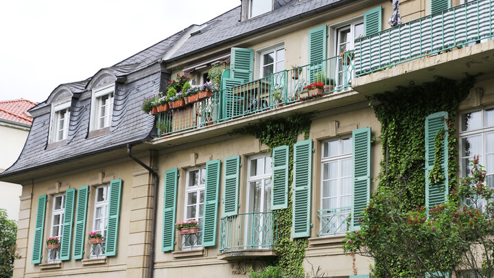 Beige house facade with light green open shutters. The house is partly overgrown with ivy.