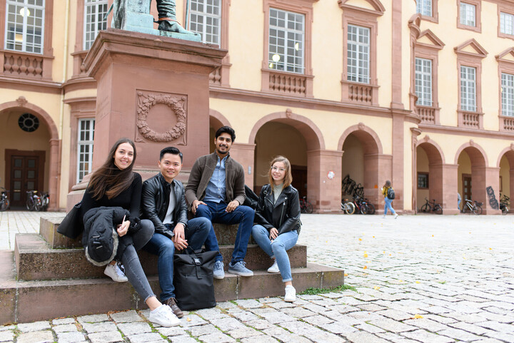 Eine Gruppe Studierende sitzt im Ehrenhof des Schlosses auf den Stufen eines Denkmals.