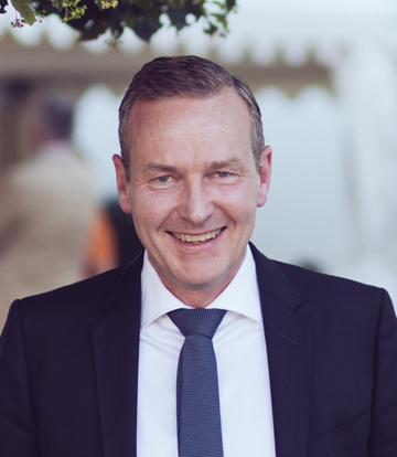 Hans Peter Grüner hat kurze graue Haare. Er trägt einen dunklen Anzug und lächelt in die Kamera.