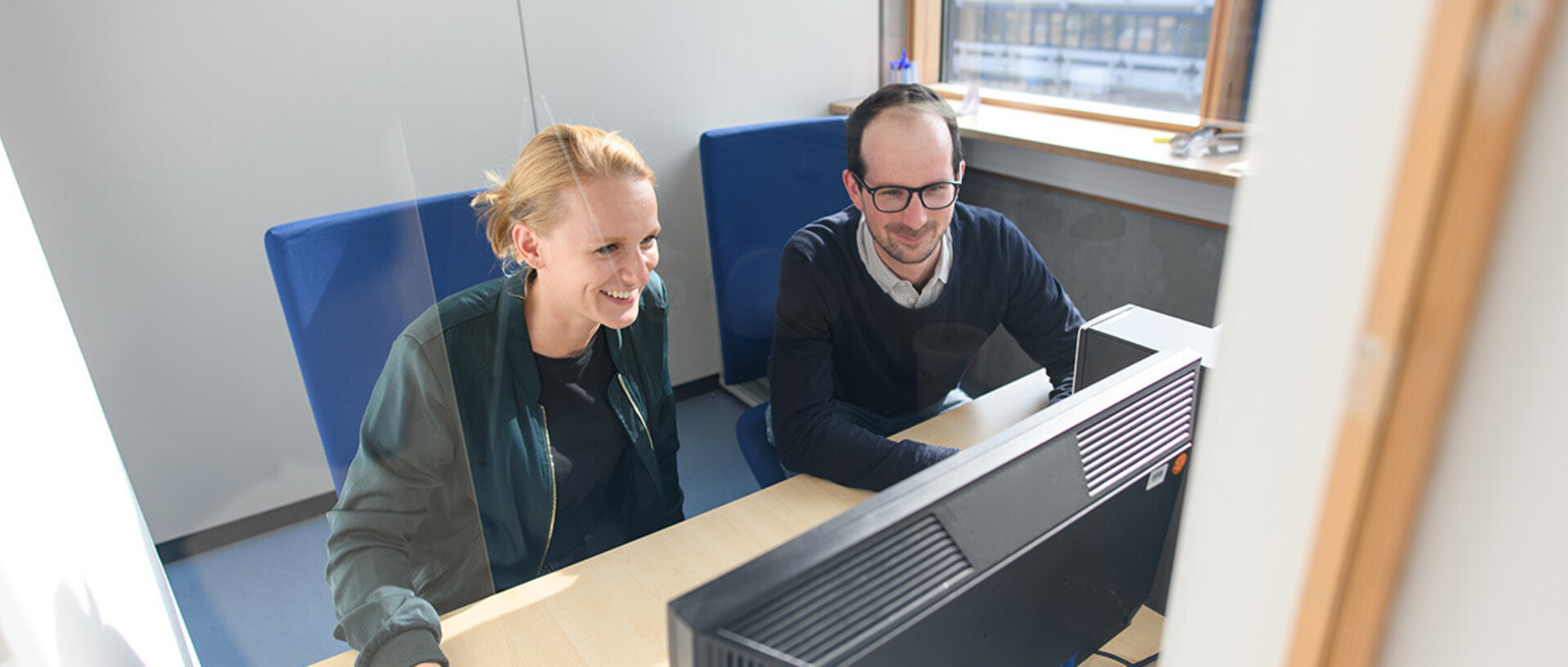 Eine Frau und ein Mann sitzen nebeneinander vor einem Computerbildschirm und blicken lächelnd darauf. Der Raum im Hintergrund hat große Fenster, durch die Tageslicht einfällt.