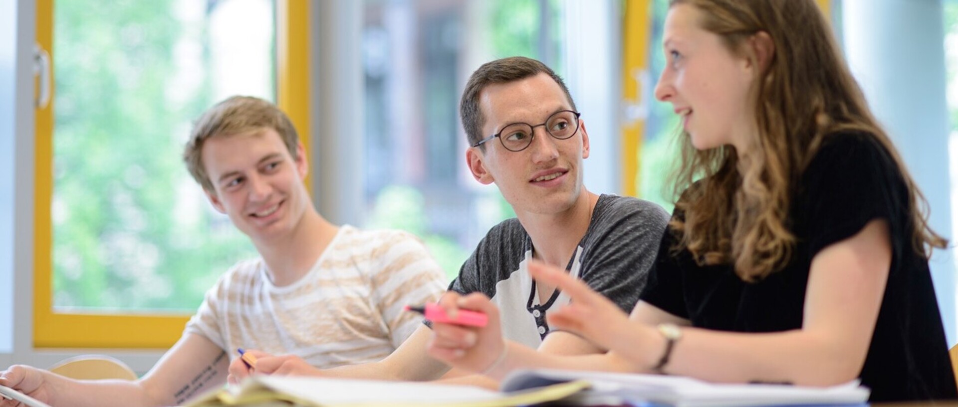 Drei Studierende in einem Übungsraum. Vor ihnen befinden sich Lernmaterialien. Eine Studentin spricht, sie trägt ein schwarzes T-Shirt und hat mittellange braune Haare, in ihrer Hand hält sie einen pinken Textmarker. Die Studenten neben ihr schauen sie an. Der Student in der Mitte hat kurze braune Haare und trägt eine Brille. Er trägt ein weißes Shirt mit grauen Ärmeln. Der Student auf der linken Seite hat blonde kurze Haare und trägt ein beige-weiß gestreiftes Shirt.
