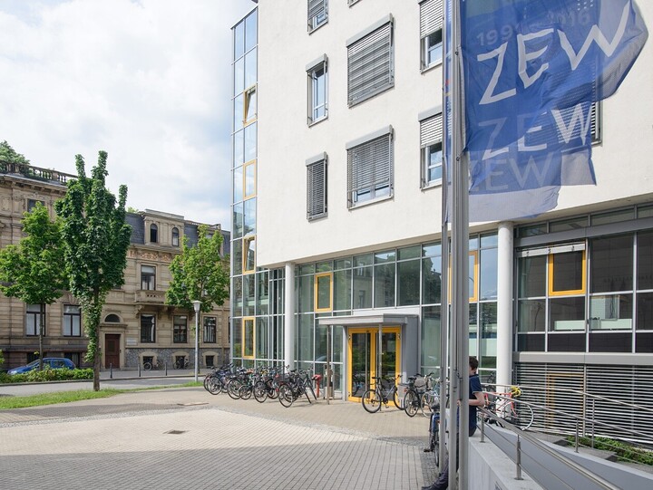 Blick auf das Abteilungsgebäude der Abteilung Volkswirtschaftslehre. Auf der linken Bildseite stehen Bäume vor anderen Gebäude der Universität im Block L5. Rechtsseitig befindet sich das VWL-Gebäude vor dem einige Fahrräder stehen. In der rechten oberen Ecke wehen die dunkelblauen Fahnen des ZEW.