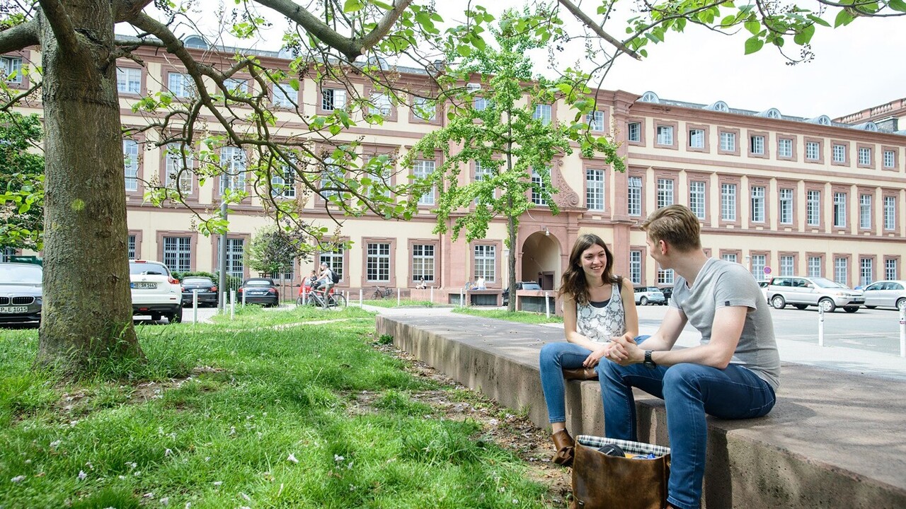 Zwei Studierende sitzen auf einer Einfassung vor dem Westflügel der Universität Mannheim und unterhalten sich.