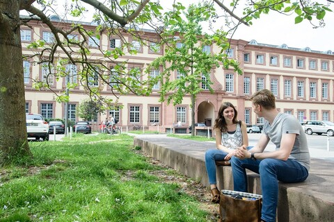 Zwei Studierende sitzen auf einer Einfassung vor dem Westflügel der Universität Mannheim und unterhalten sich.