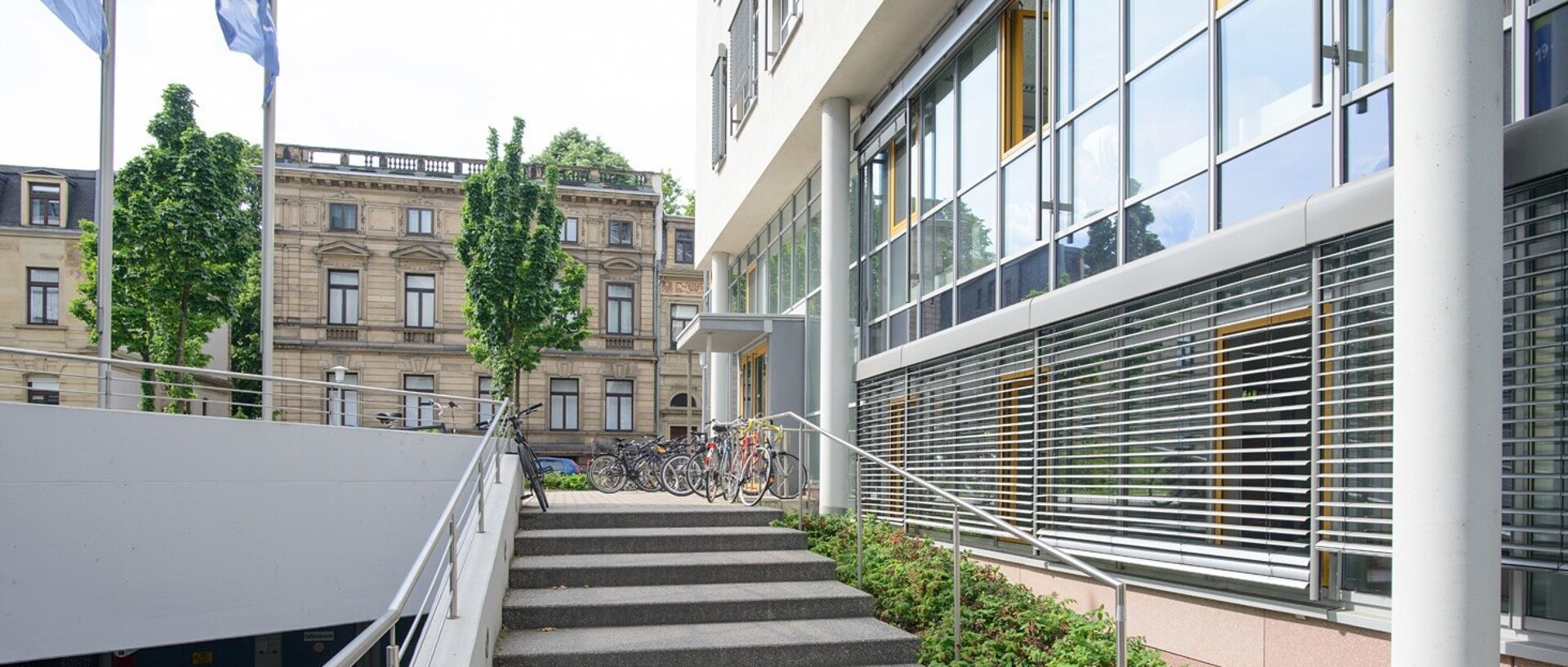 Blick auf das Gebäude der Abteilung Volkswirtschaftslehre. Auf der linken Bildseite befindet sich ein weiteres Universitätsgebäude im Block L5, vor welchem Bäume stehen. Mittig führt eine Treppe zum Eingang in das VWL-Gebäude. Im unteren Stock des Gebäudes sind die Jalousien heruntergefahren.