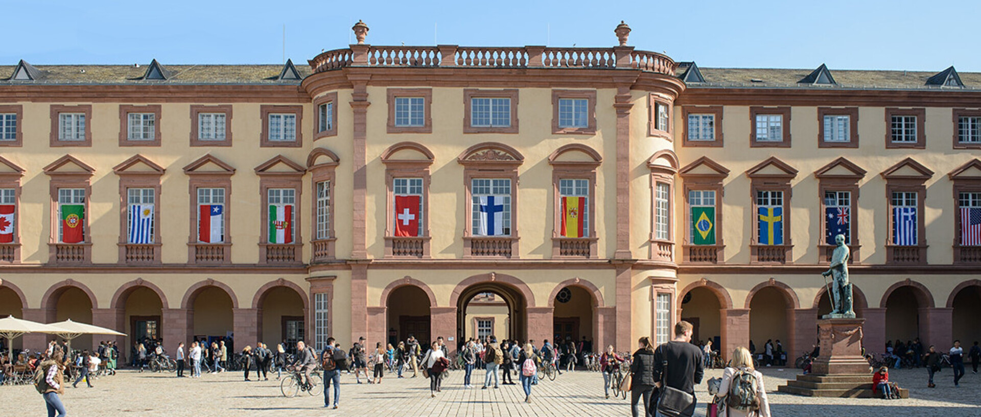 Ostflügel der Universität Mannheim mit vielen Flaggen verschiedener Länder an den Fenstern im ersten Stock. Über den Ehrenhof laufen viele Menschen