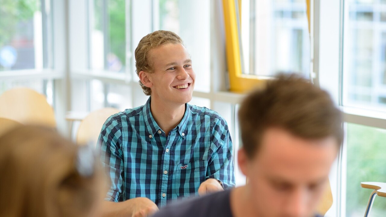 Im Zentrum des Bildes sitzt ein lächelnder Student. Er ist blond und trägt ein blau kariertes Hemd. Hinter ihm erkennt man eine Glasfassade, vor ihm sitzen weitere Studierende.