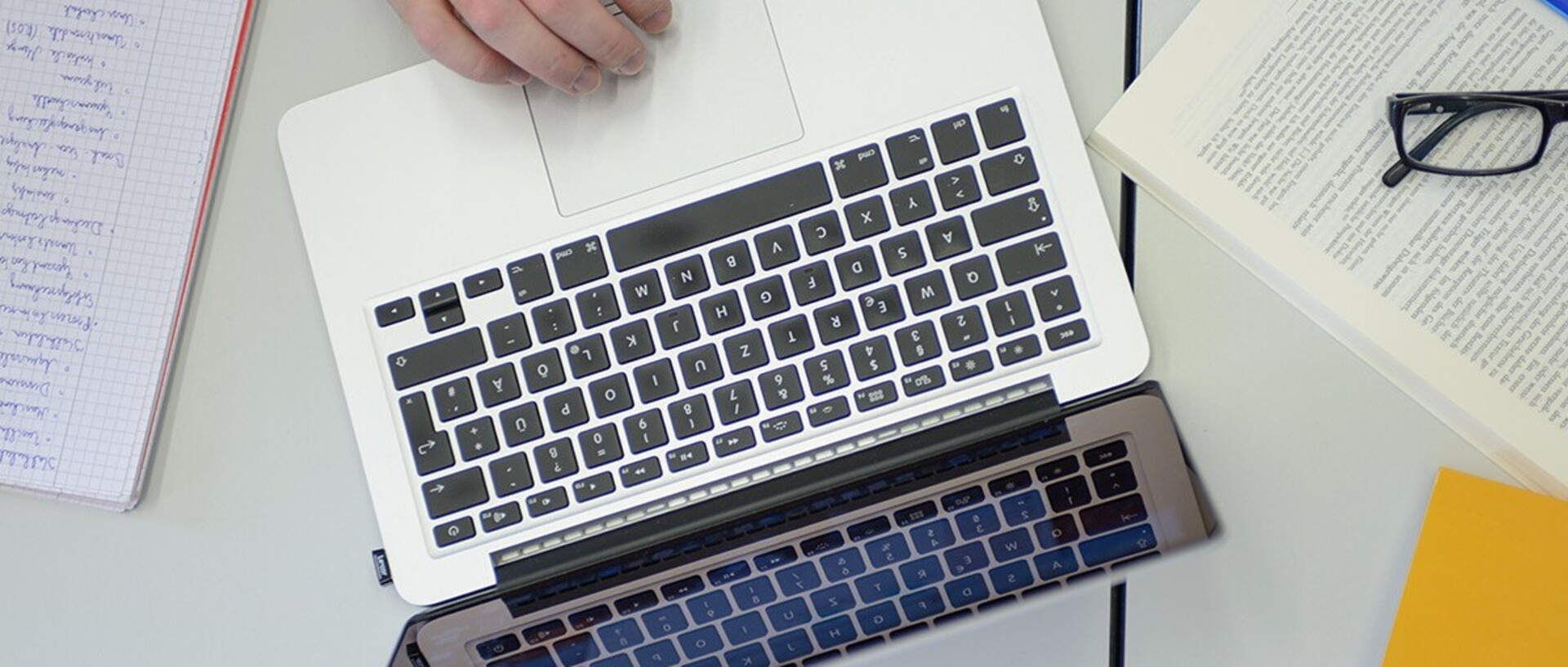 An open laptop stands on a table. Around it is a written pad on the left side of the picture and on the right side an open book with a pair of glasses with black frames and a yellow piece of paper.