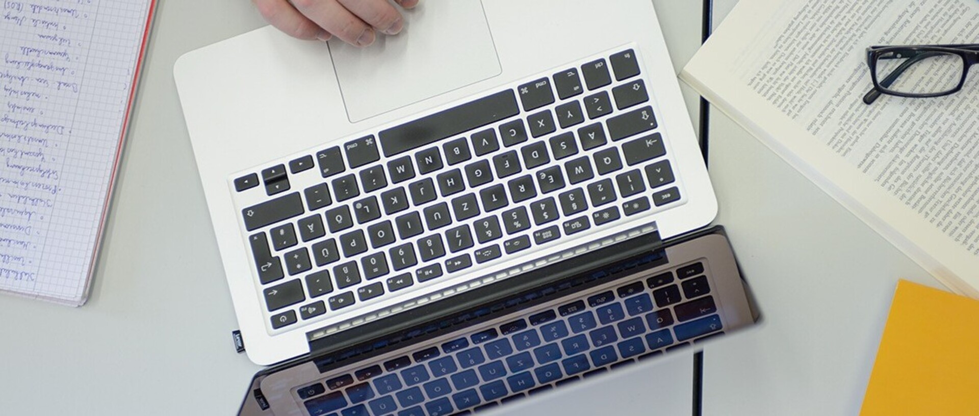 Ein aufgeklappter Laptop steht auf einem Tisch. Darum herum liegen ein beschriebener Block auf der linken Bildseite und auf der rechten Bildseite ein aufgeschlagenes Buch, auf dem eine Brille mit schwarzer Fassung liegt, sowie ein gelber Zettel.