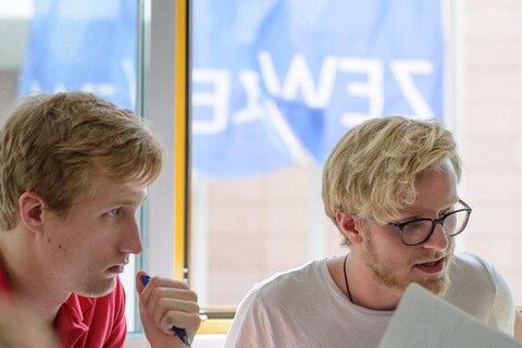 Zwei Studenten sitzen in einem Übungsraum und arbeiten. Im Hintergrund wehen die dunkelblauen Fahnen des ZEW.