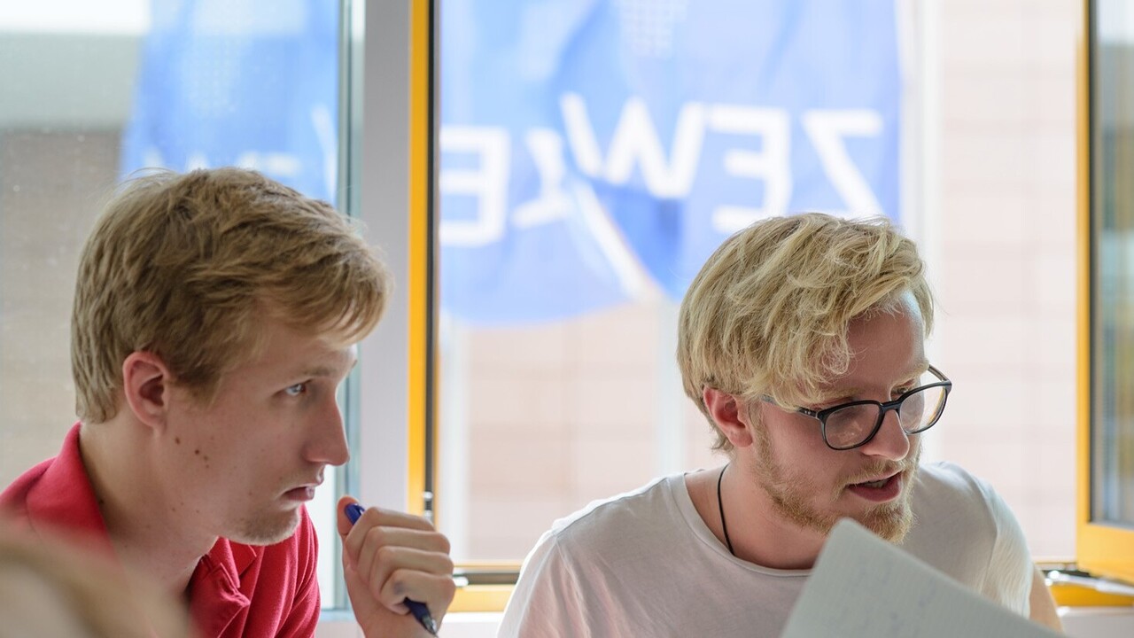 Zwei Studenten sitzen in einem Übungsraum und arbeiten. Im Hintergrund wehen die dunkelblauen Fahnen des ZEW.