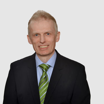 Prof. Maug hat kurze graue Haare und trägt ein dunkelblaues Jacket mir hellblauem Hemd und grüner Krawatte.