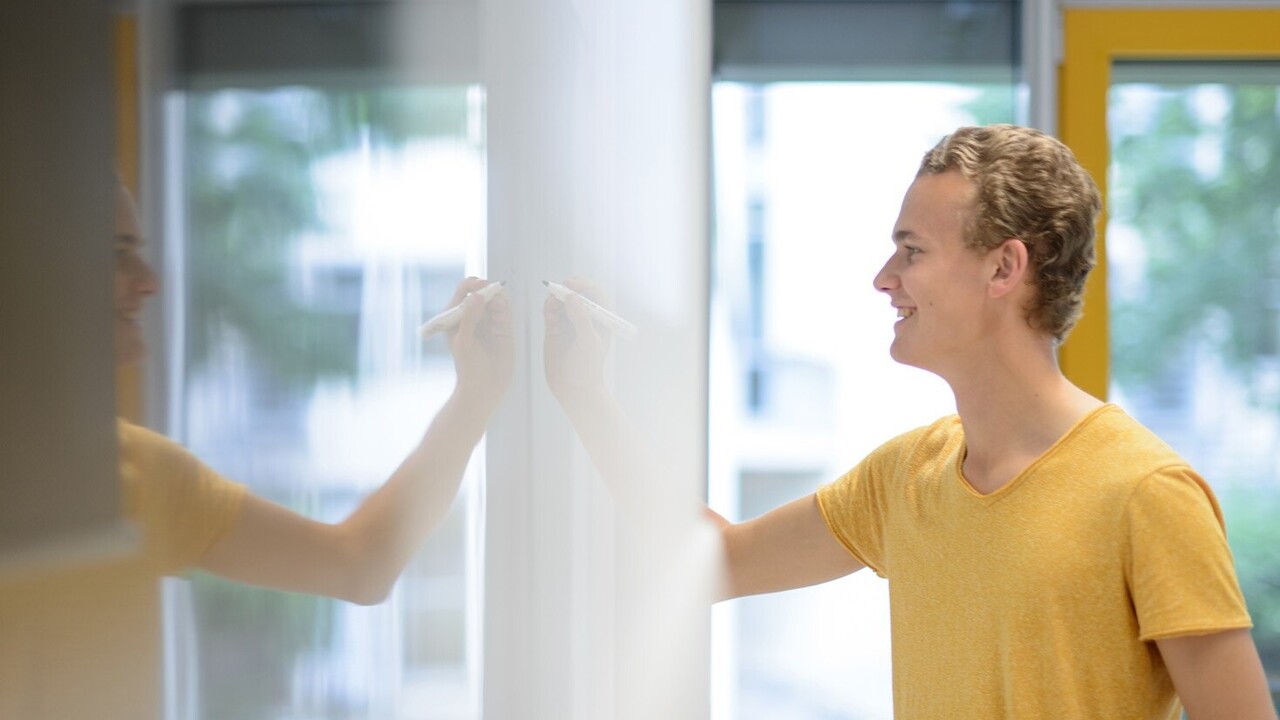 Ein Student mit blonden Haaren beschreibt ein Whiteboard. Er trägt ein gelbes T-Shirt und lächelt. Das Whiteboard befindet sich auf der linken Bildseite, der Student auf der rechten.
