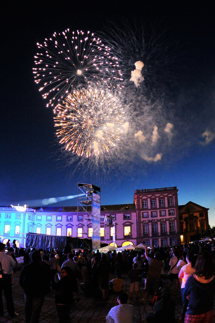 Eine Veranstaltung bei Nacht auf dem Ehrenhof. Viele Menschen Blicken in den Himmel, das Schloss wird blau, lila und rosa beleuchtet. Am Nachthimmel explodieren Feuerwerksraketen.