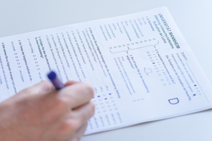 Ein Fragebogen zur Lehrevaluation wird mit einem blauen Stift ausgefüllt. Die Hand der Person, welche den Bogen ausfüllt, befindet sich am rechten Bildrand.