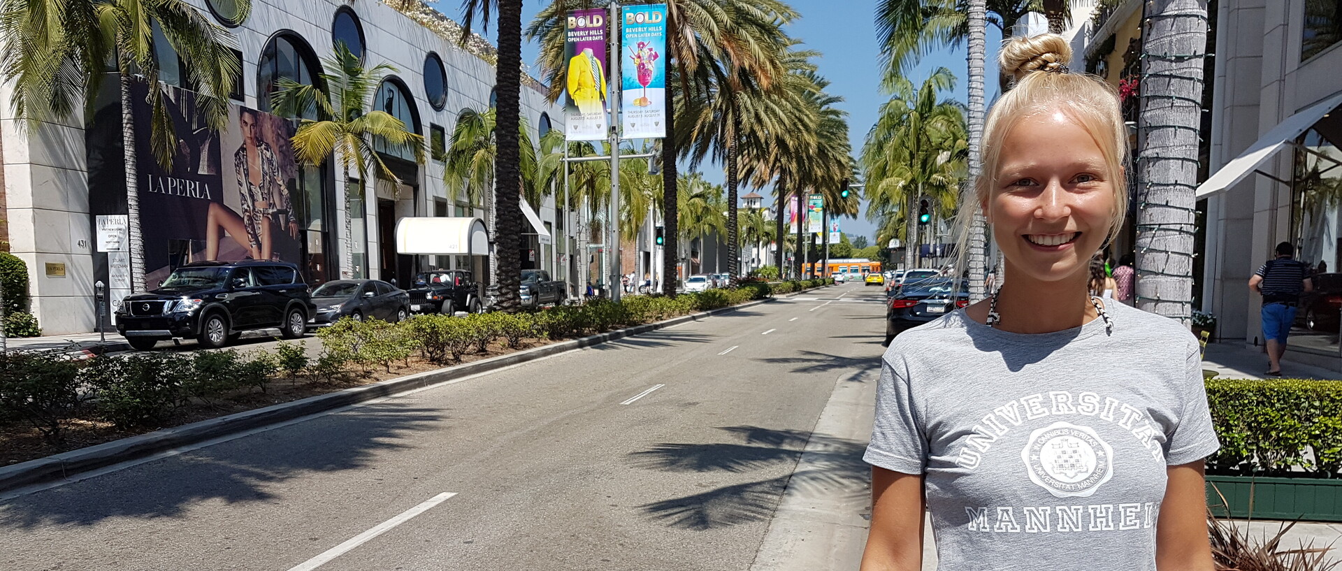 Studentin mit blonden Haaren und grauem Uni Mannheim Shirt und Jeans-Shorts. Sie steht auf einer Straße in Beverly Hills mit Palmen.
