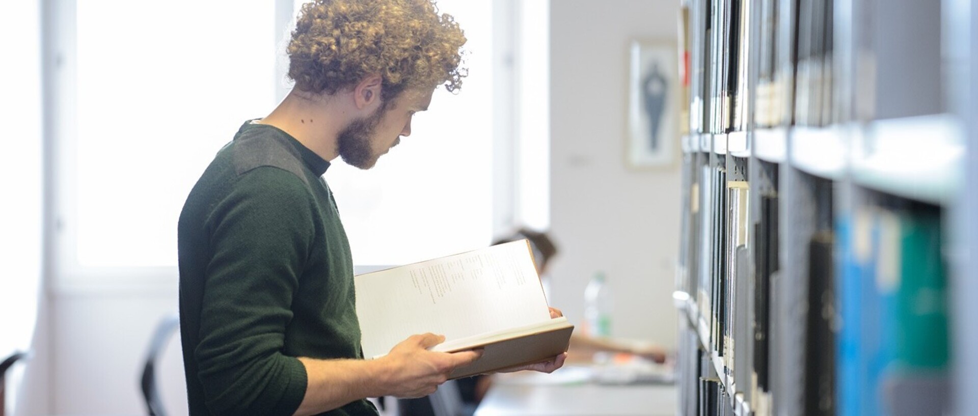 A student with blond curls and a green top is standing in front of a bookshelf. He is reading an open book.
