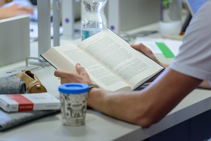 Eine Person liest ein Buch an einem weißen, mit weiteren Gegenständen belegten Tisch.