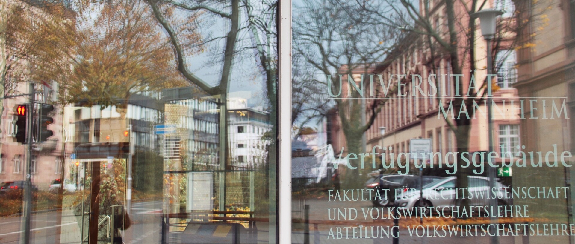 Spiegelung in der Glasfassade des Gebäudes der Abteilung Volkswirtschaftslehre. Auf der rechten Glasscheibe steht: Universität Mannheim; Verfügungsgebäude; Fakultät für Rechtswissenschaft und Volkswirtschaftslehre; Abteilungsgebäude Volkswirtschaftslehre.